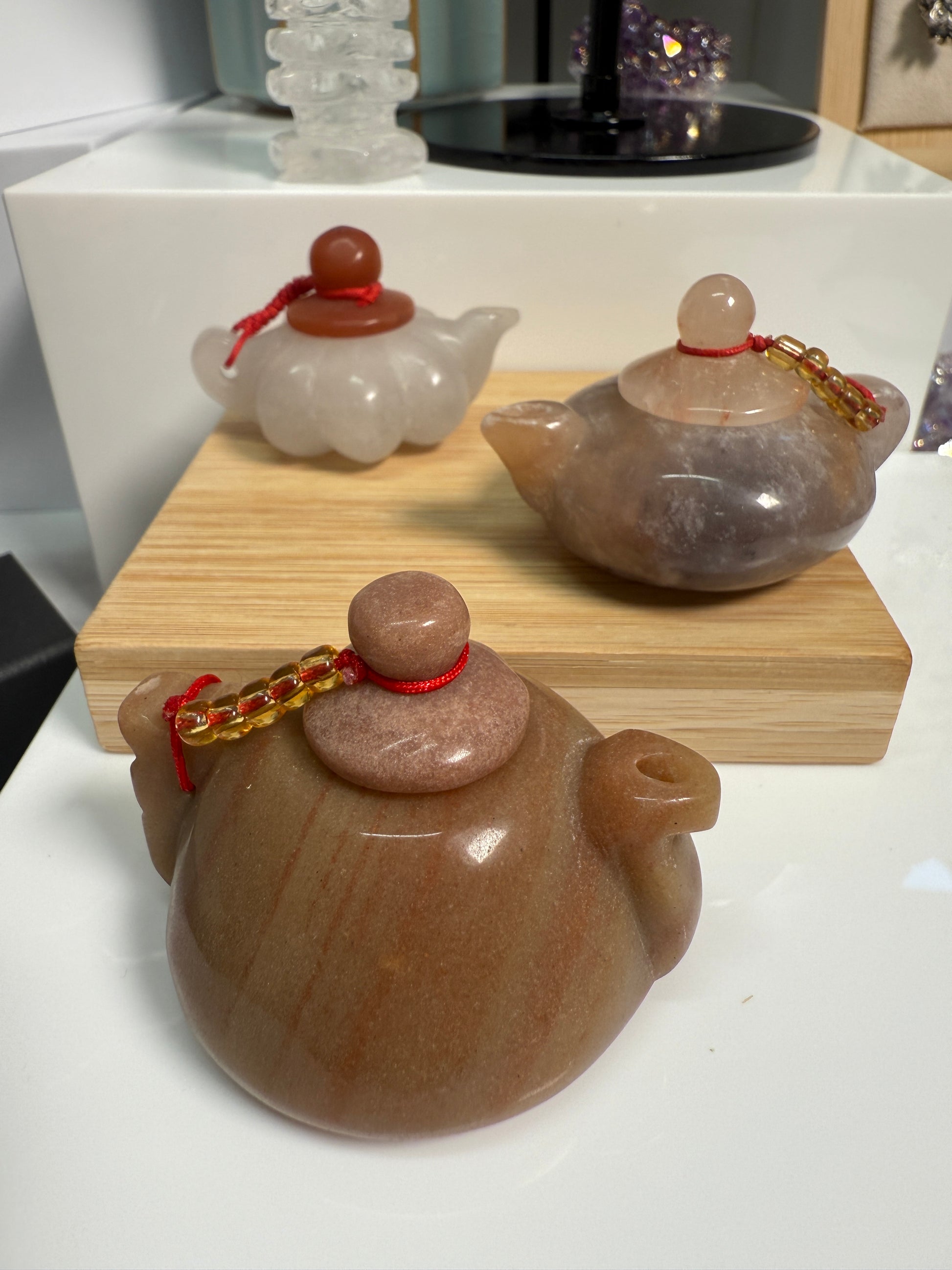Image of three mini crystal teapots displayed —White Jade, Golden Jasper, and Purple Jade—each polished and carved in a classic teapot shape. The 2-inch figurines are arranged on a flat surface as elegant decorative accents.