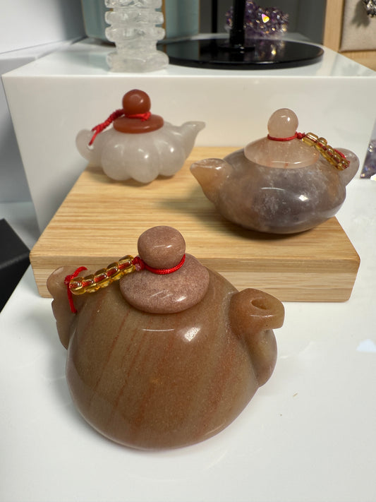 Image of three mini crystal teapots displayed —White Jade, Golden Jasper, and Purple Jade—each polished and carved in a classic teapot shape. The 2-inch figurines are arranged on a flat surface as elegant decorative accents.